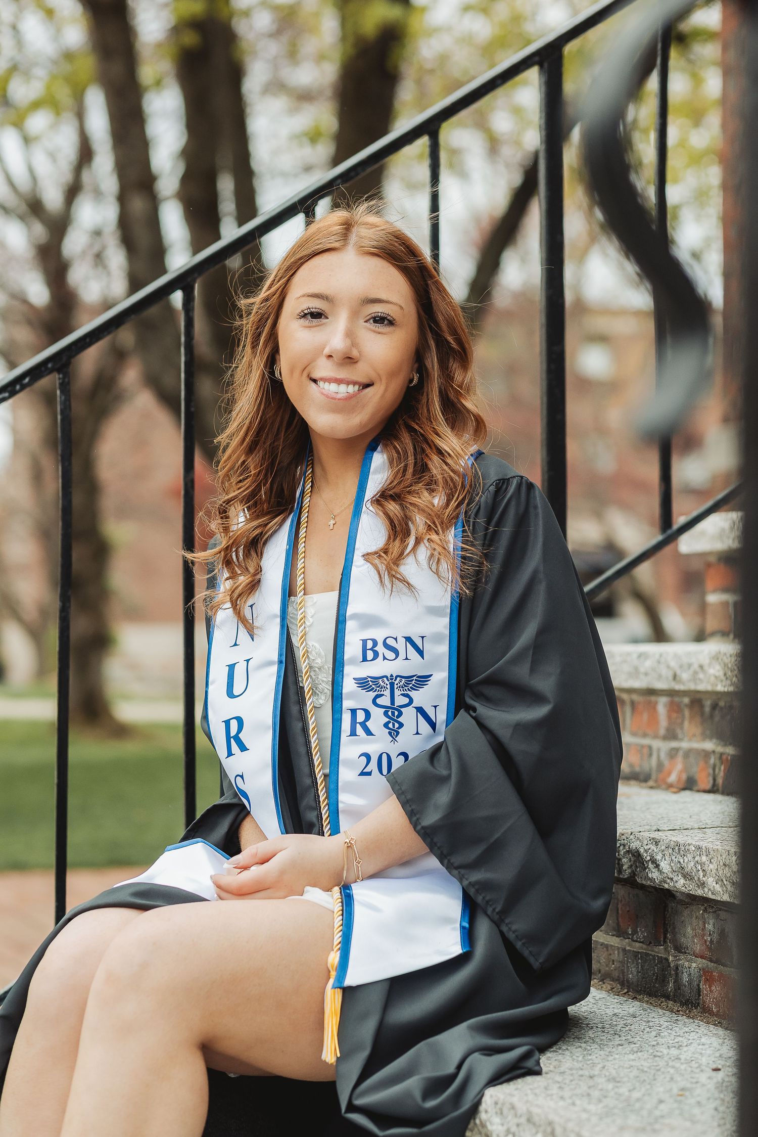Smiling female BSN nursing graduate in cap and gown sitting on steps outdoors during graduation celebration.