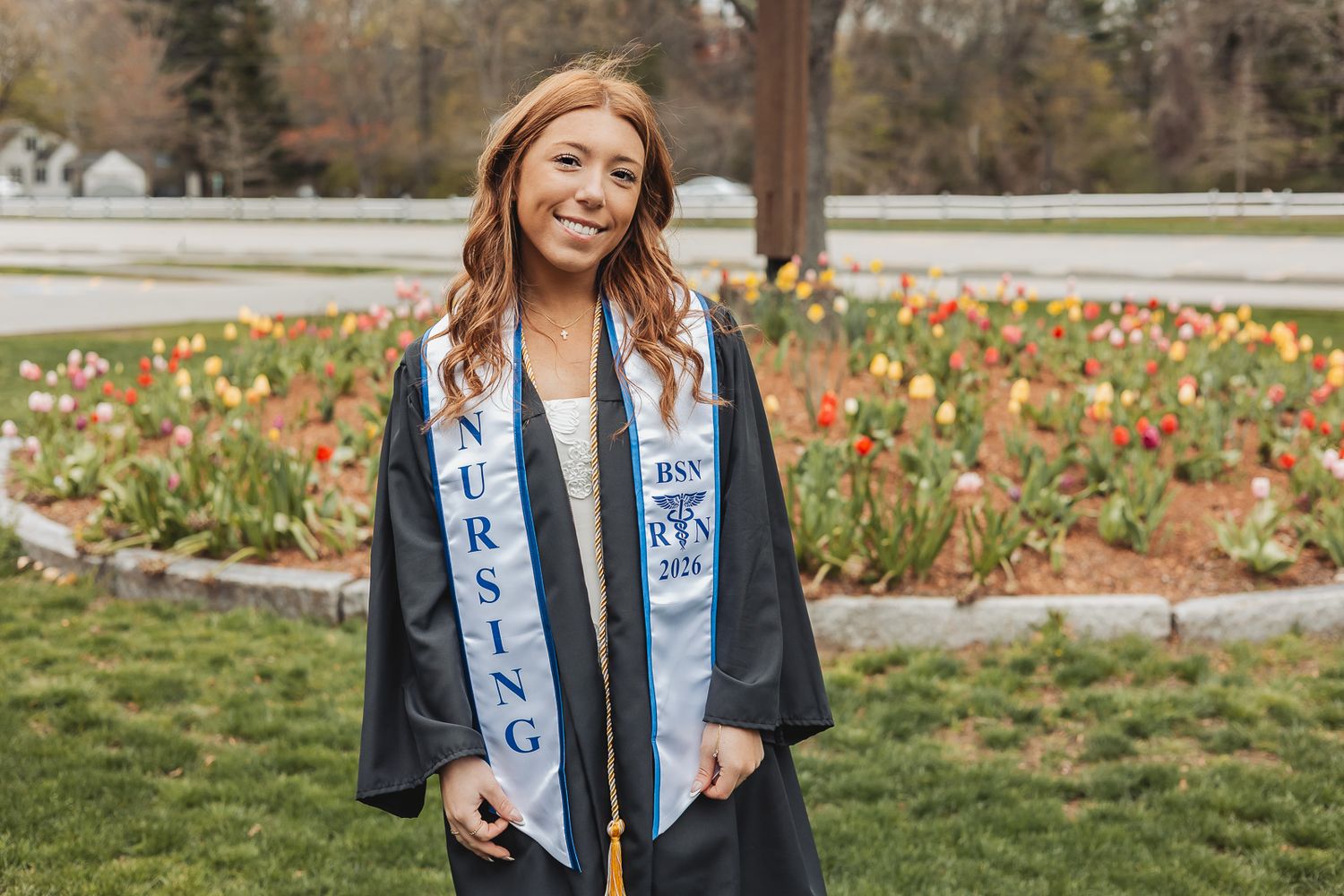 Smiling nursing graduate in black cap and gown with blue BSN nursing sash stands outdoors near colorful tulip garden.