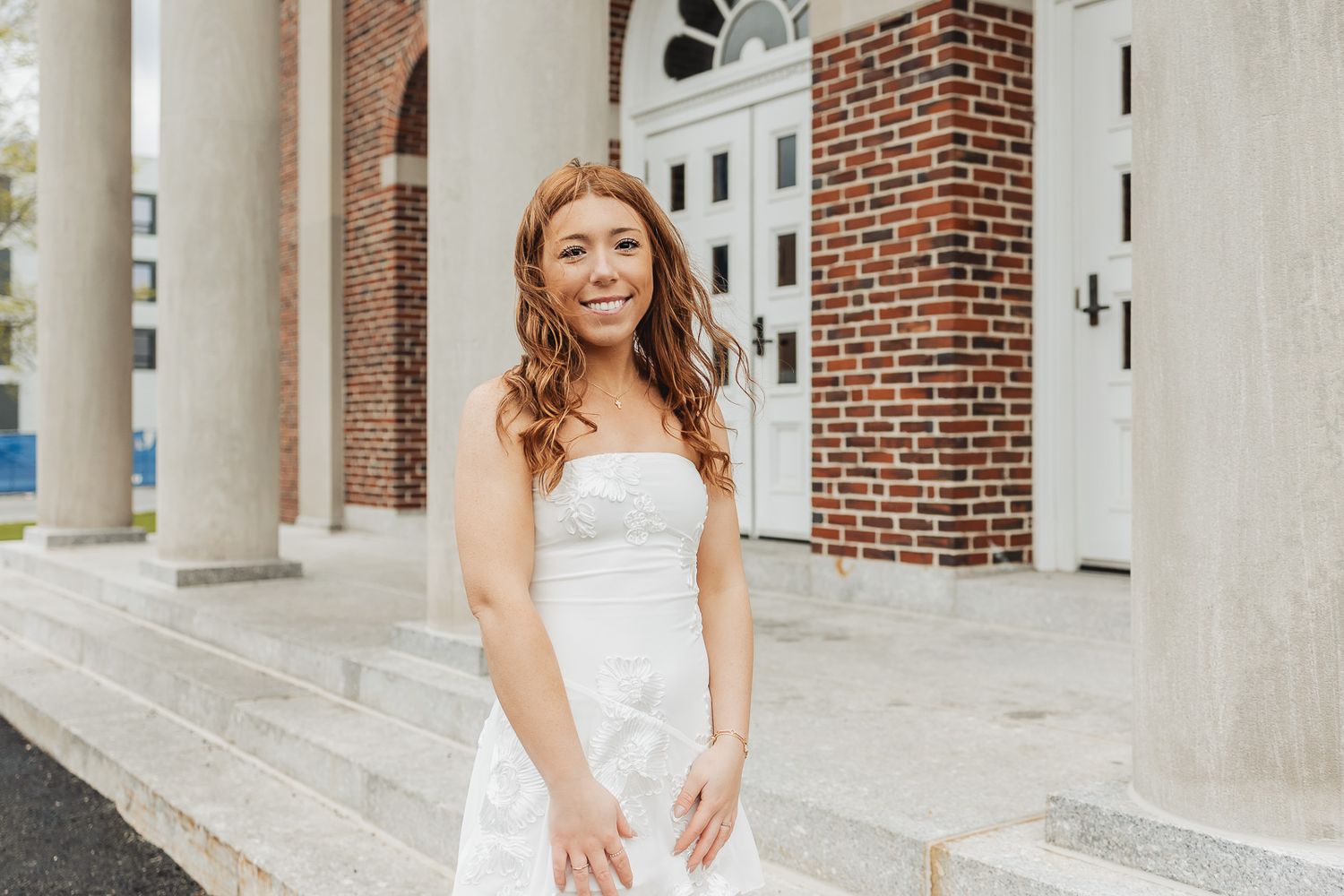 Young woman in white strapless dress smiling on steps of brick building with white columns.