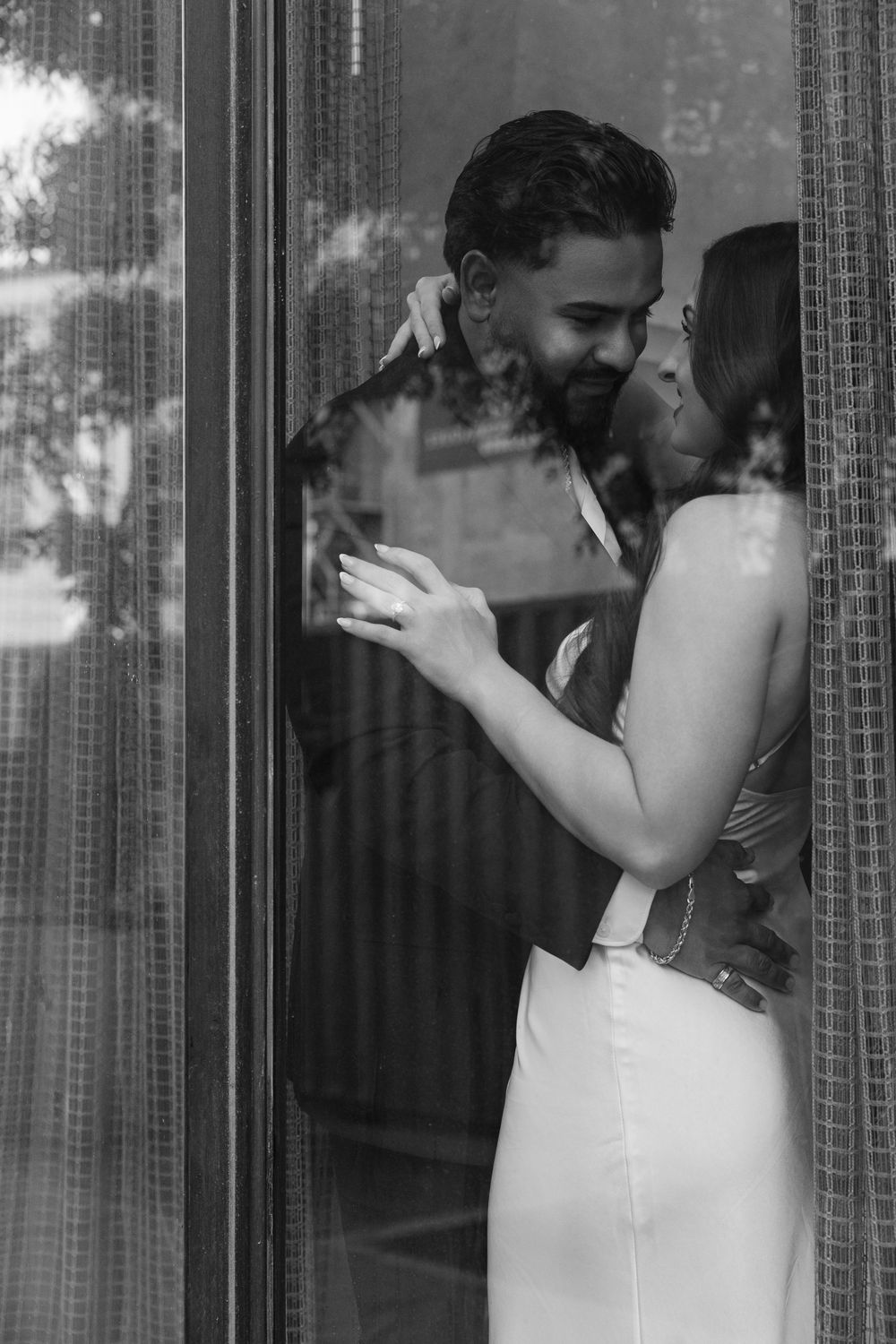 Black and white photo of a couple embracing and smiling at each other by a glass door in elegant attire.