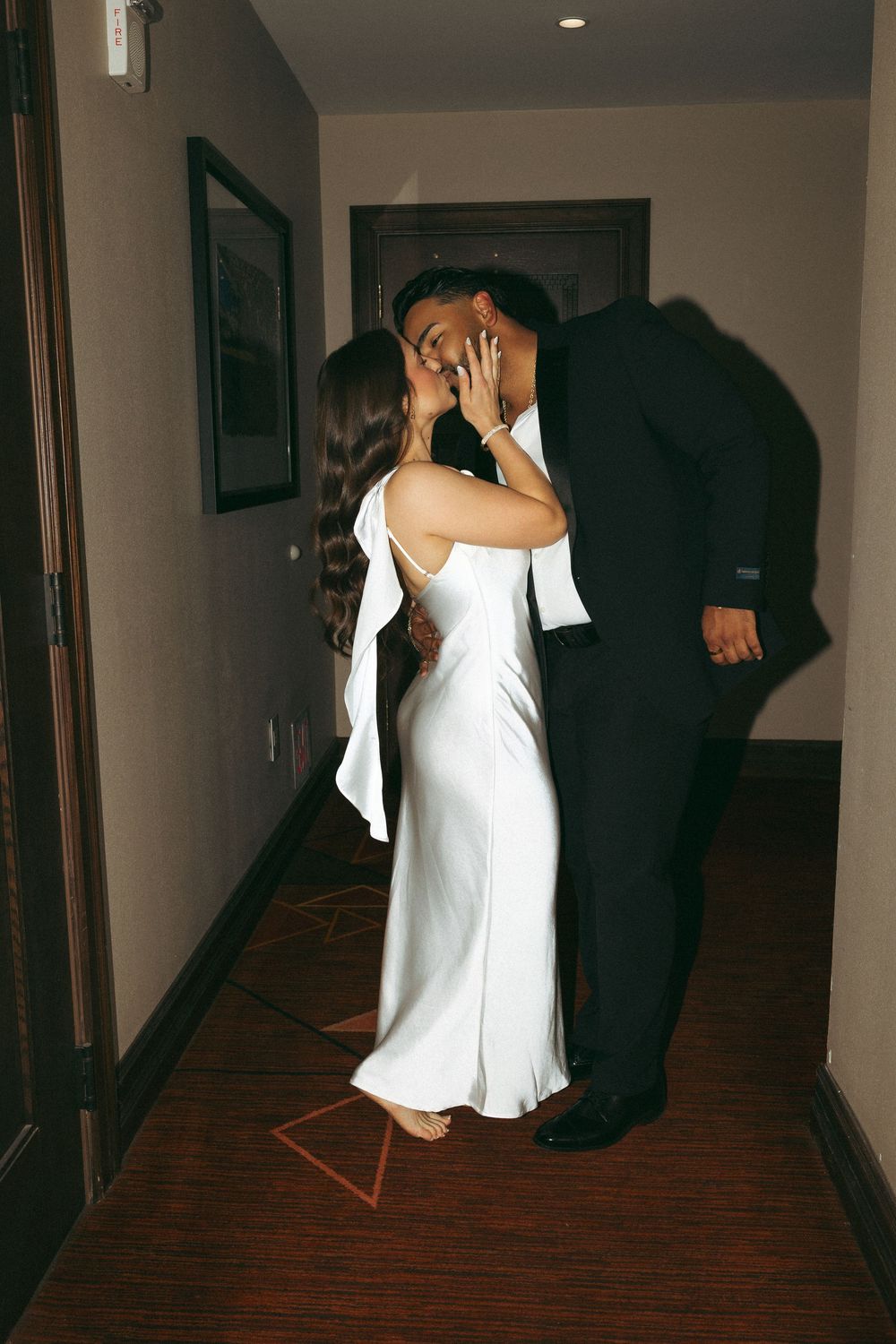 Couple sharing a romantic kiss in a hotel hallway, bride in white satin dress and groom in black suit.