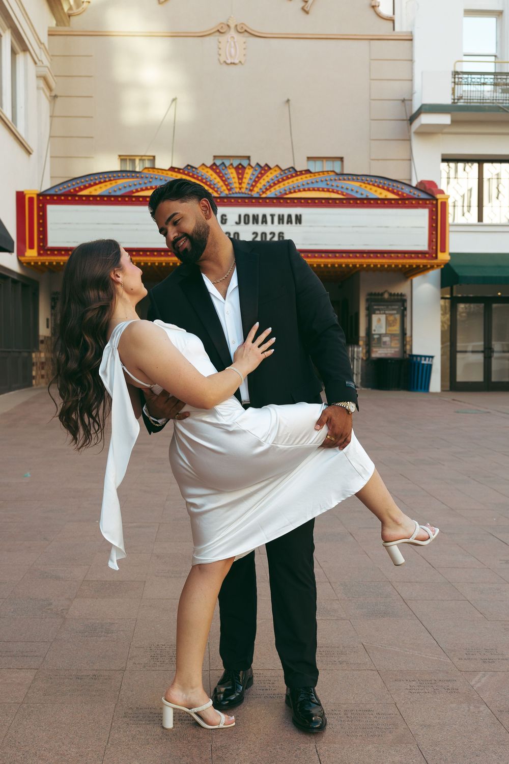 Groom lifting bride in white dress outside a theater marquee, sharing a joyful dance together.