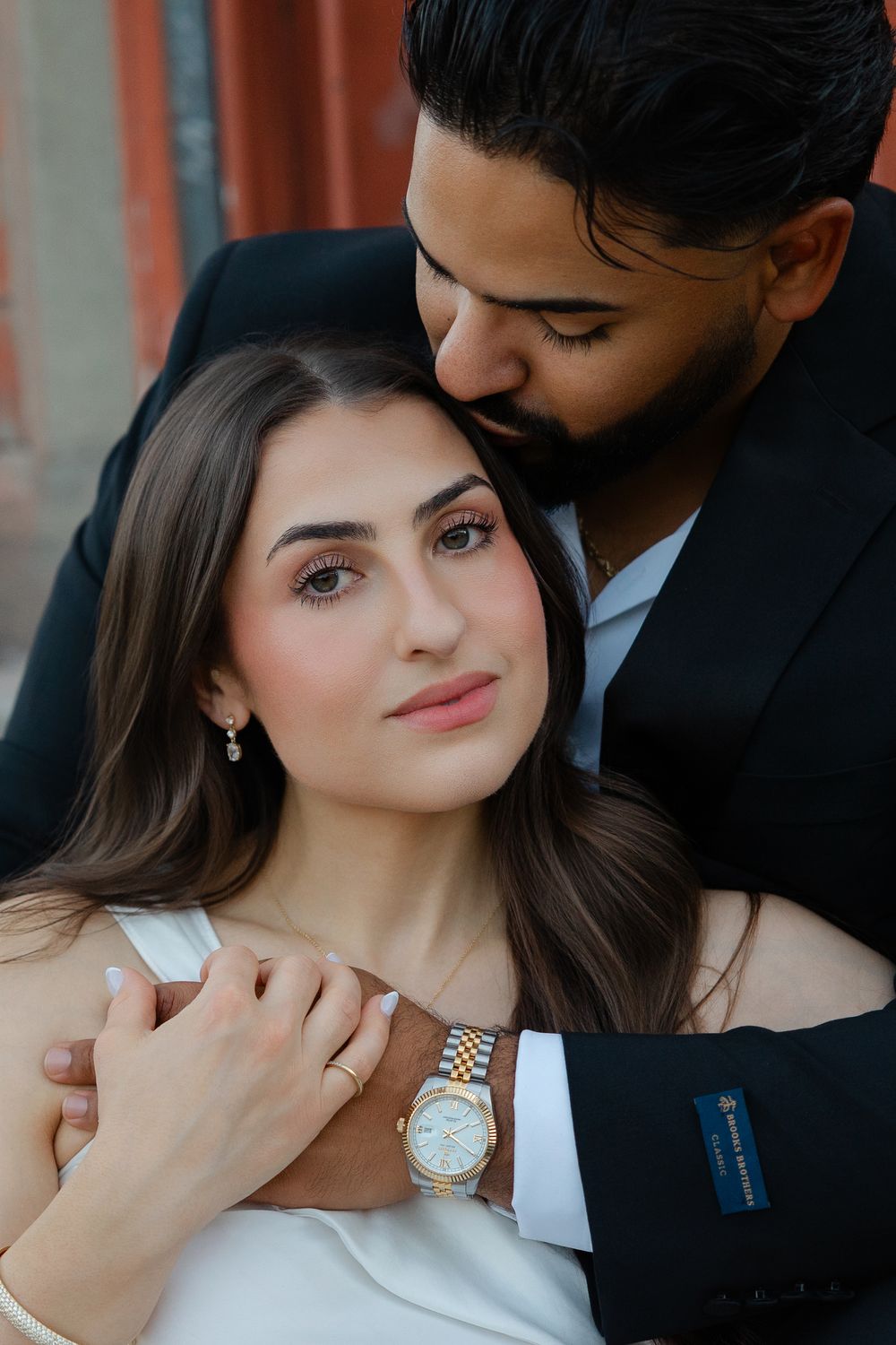 Romantic couple portrait with man in black suit embracing woman in white dress, wearing a gold Rolex watch.