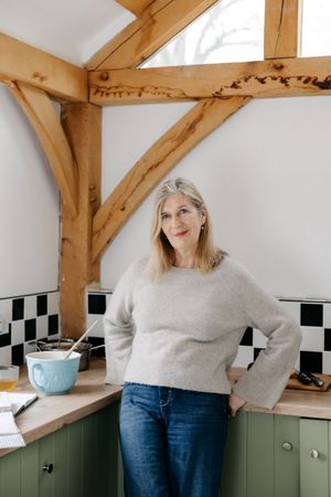 Woman in gray sweater and jeans smiling in a rustic kitchen with green cabinets and wooden beam accents.
