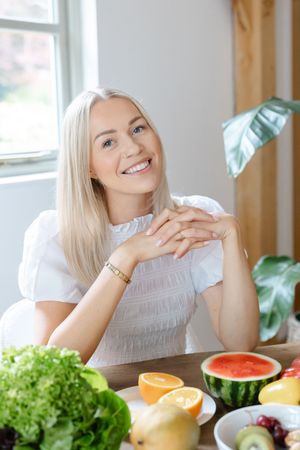 Smiling blonde woman sitting at a table with fresh fruits and vegetables, wearing a white blouse in a bright kitchen.
