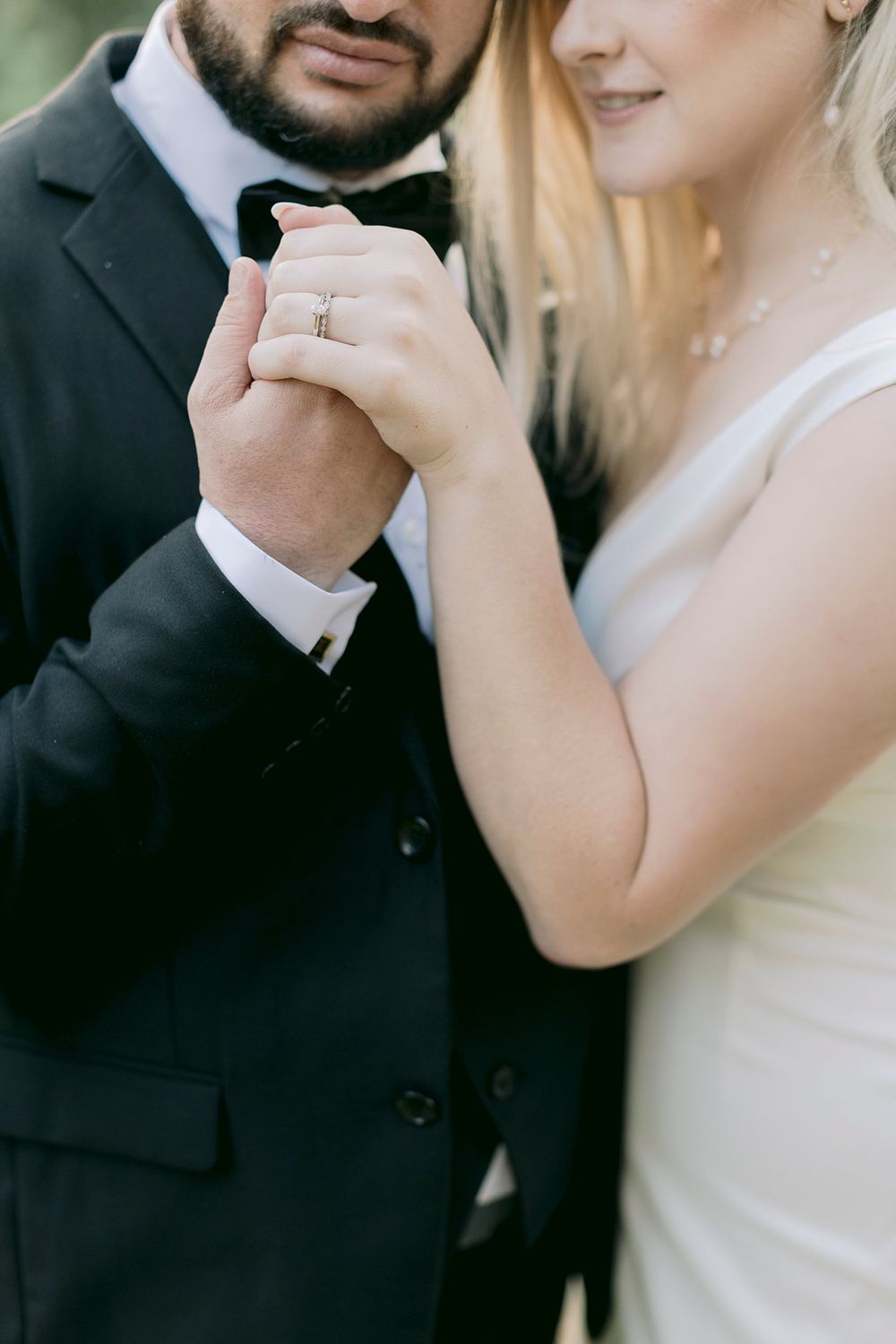 Close-up of bride and groom holding hands, showcasing diamond engagement ring, groom in black tuxedo.