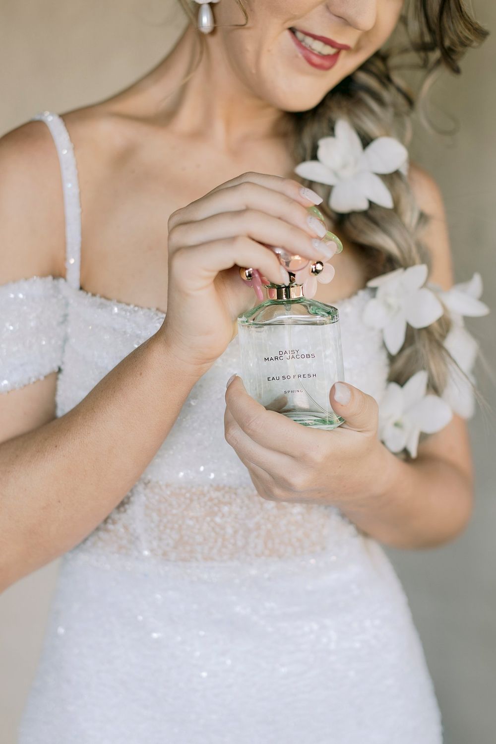 Smiling bride in sequin dress holding Marc Jacobs perfume bottle with white flowers in her hair.