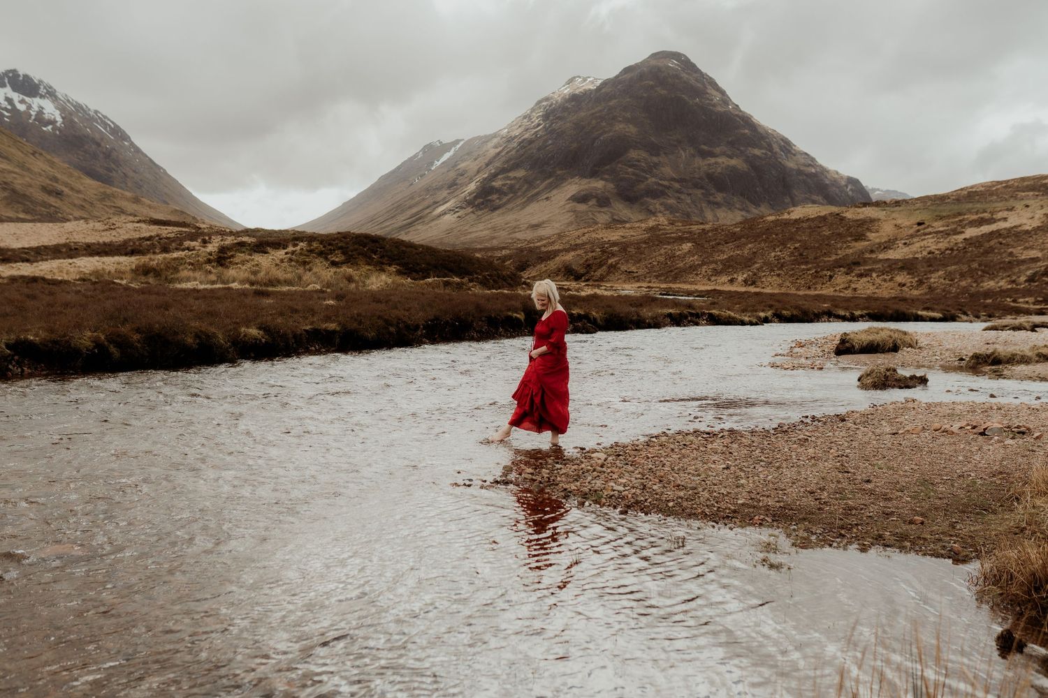 Woman in red dress wading through a river in a dramatic Scottish Highland landscape with misty mountains.
