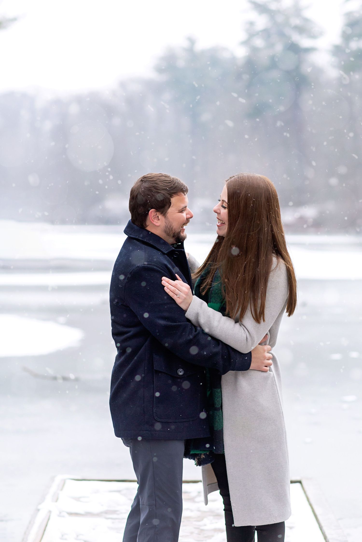 Couple embracing and smiling at each other on a snowy winter dock, with a frozen lake and trees in the background.