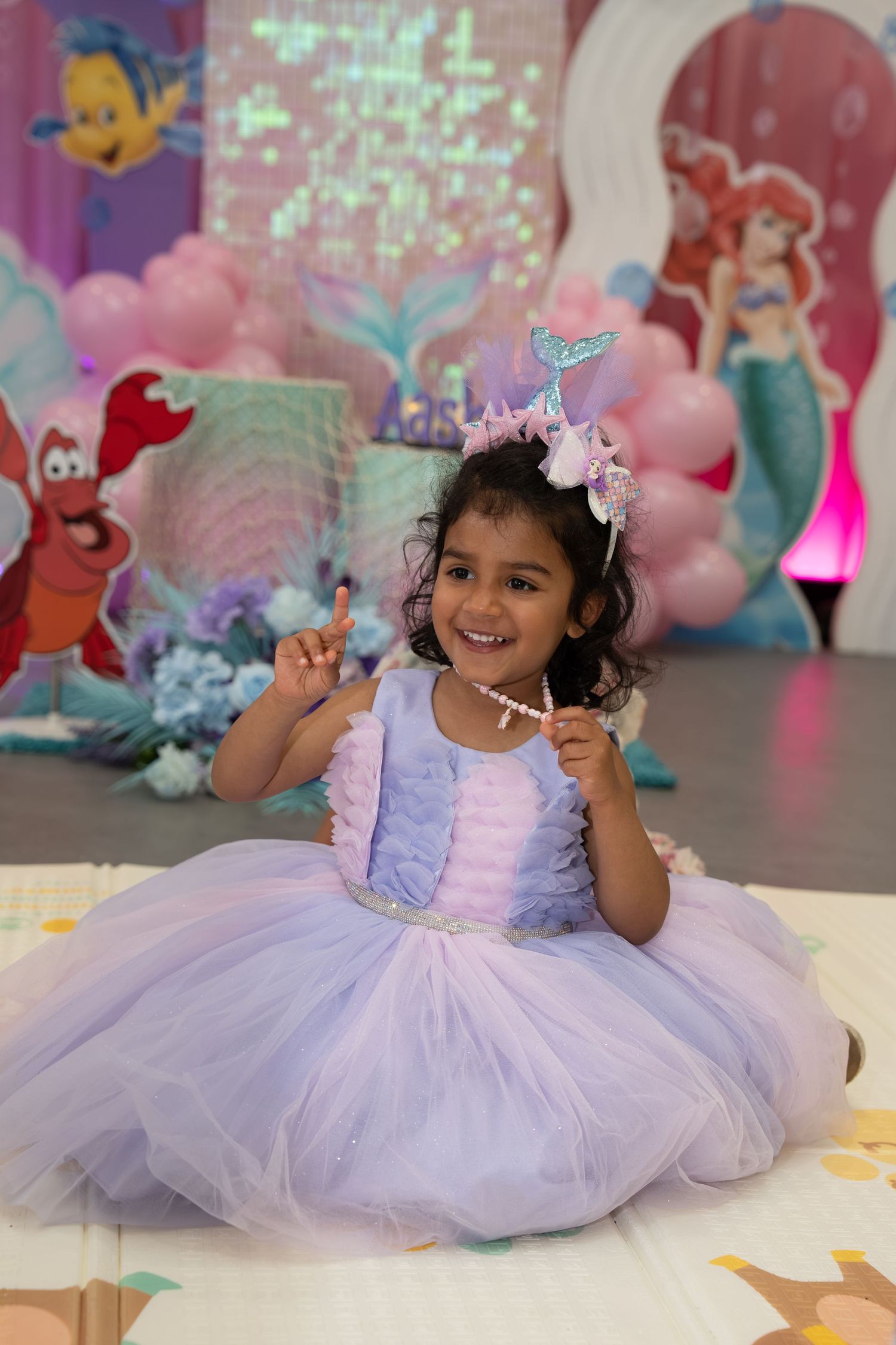 A toddler girl in a lavender tutu dress holds a wand at a Little Mermaid themed birthday party.