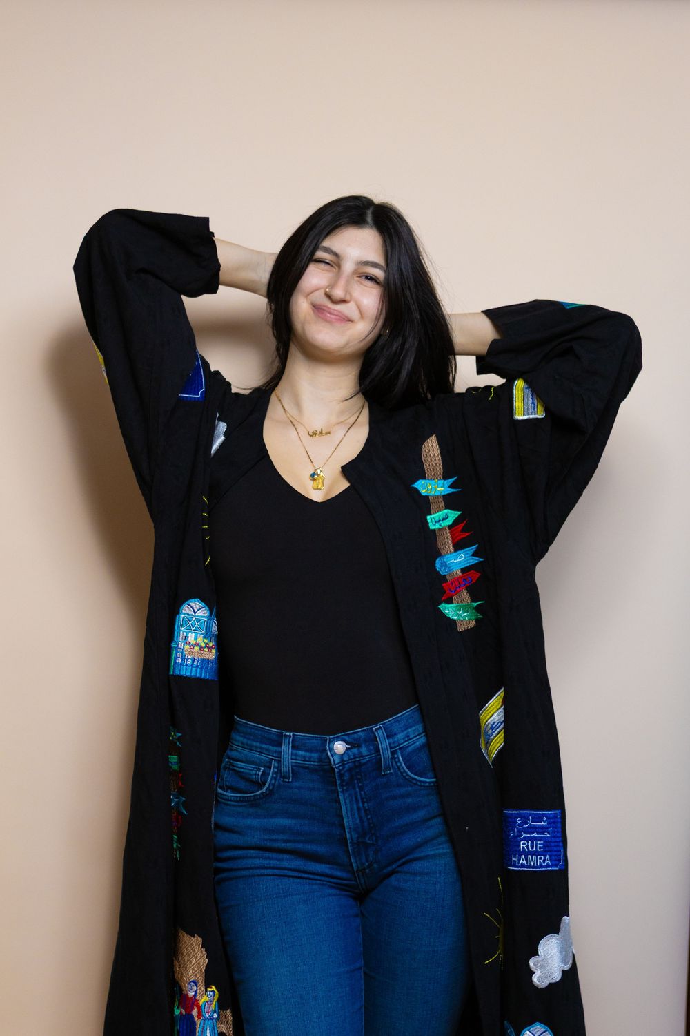Model in black cardigan with colorful patches poses casually against beige wall.