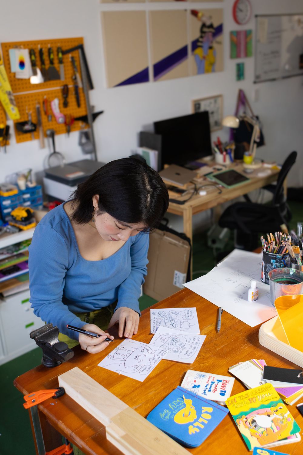 Student in blue sweater works on drawings at wooden desk with art supplies and papers spread across surface.