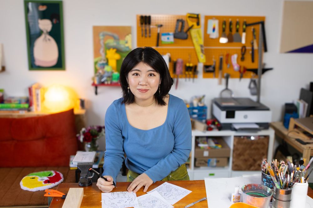 Person in blue shirt sits at wooden table writing or drawing in a well-lit room with artwork on walls.