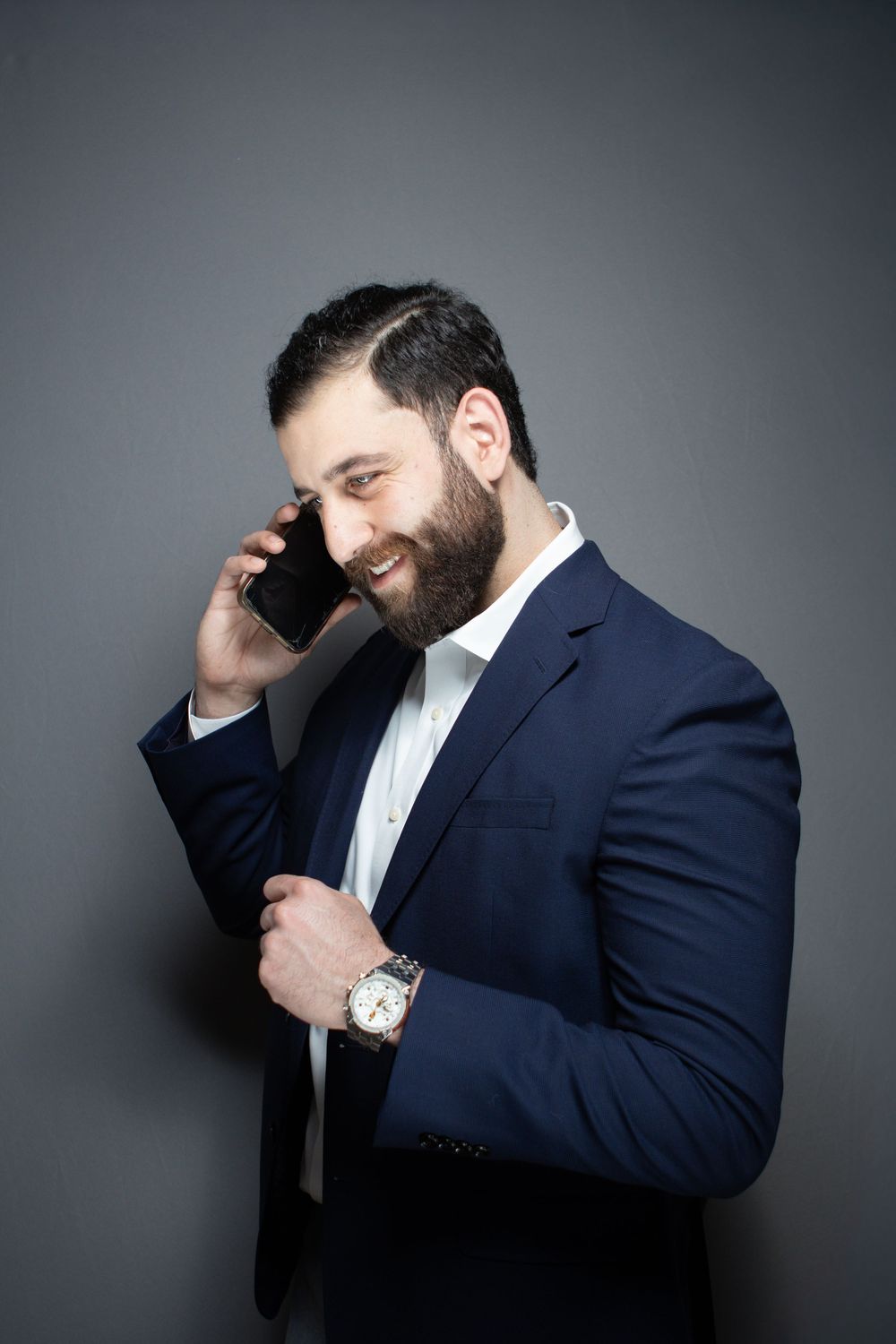 Smiling bearded businessman in navy suit talking on smartphone while checking his watch on gray background.