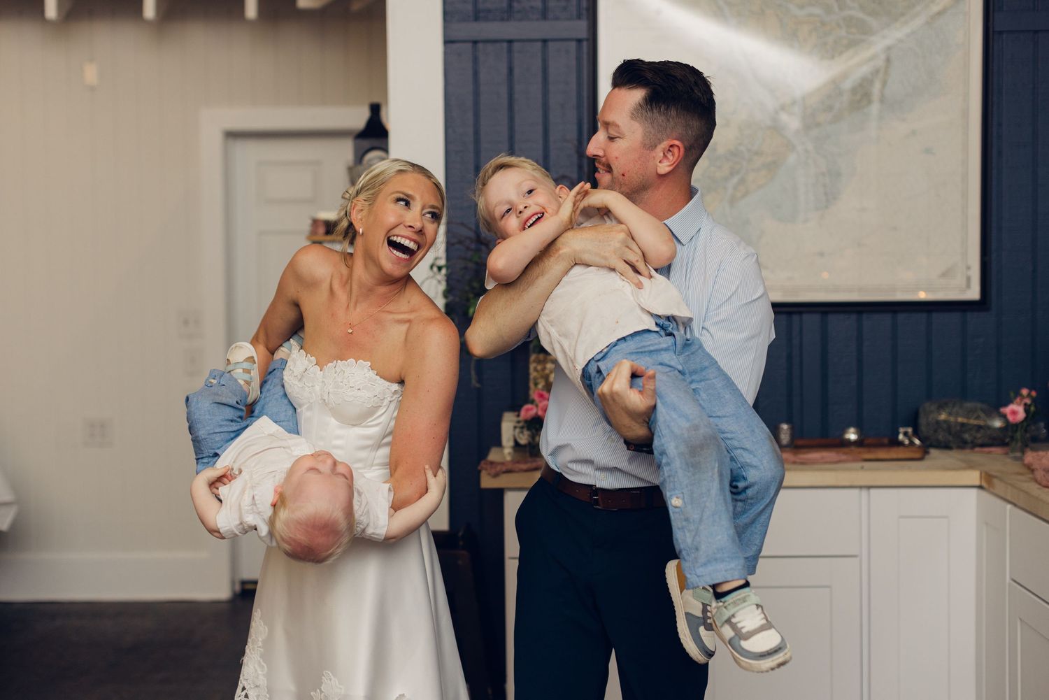 Bride and groom laughing while holding young children on their wedding day indoors.