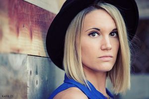 Close-up portrait of blonde woman in blue top wearing black hat with dramatic eye makeup.