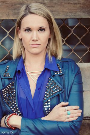 Blonde woman in blue studded jacket with arms crossed wearing colorful bracelets and turquoise ring.