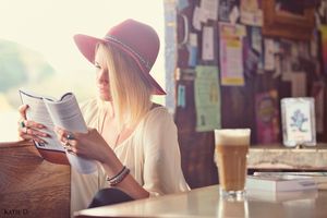 Blonde woman in red hat reading a book at a sunlit cafe table with an iced coffee drink.
