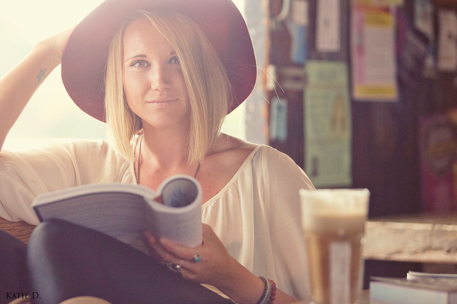 Blonde woman wearing a red hat holding a magazine while sitting in a warmly lit cafe.