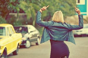 Blonde woman in blue leather jacket and black skirt gesturing with both hands near vintage cars.