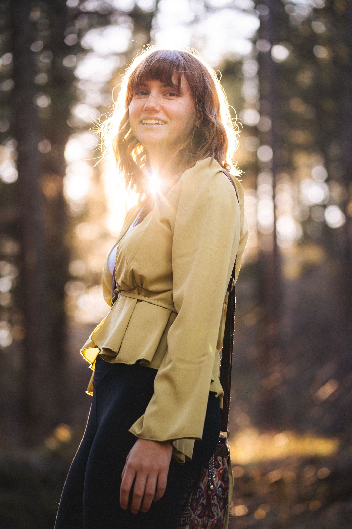 Woman in yellow blouse smiling in sunlit forest, golden backlight filtering through tall pine trees.