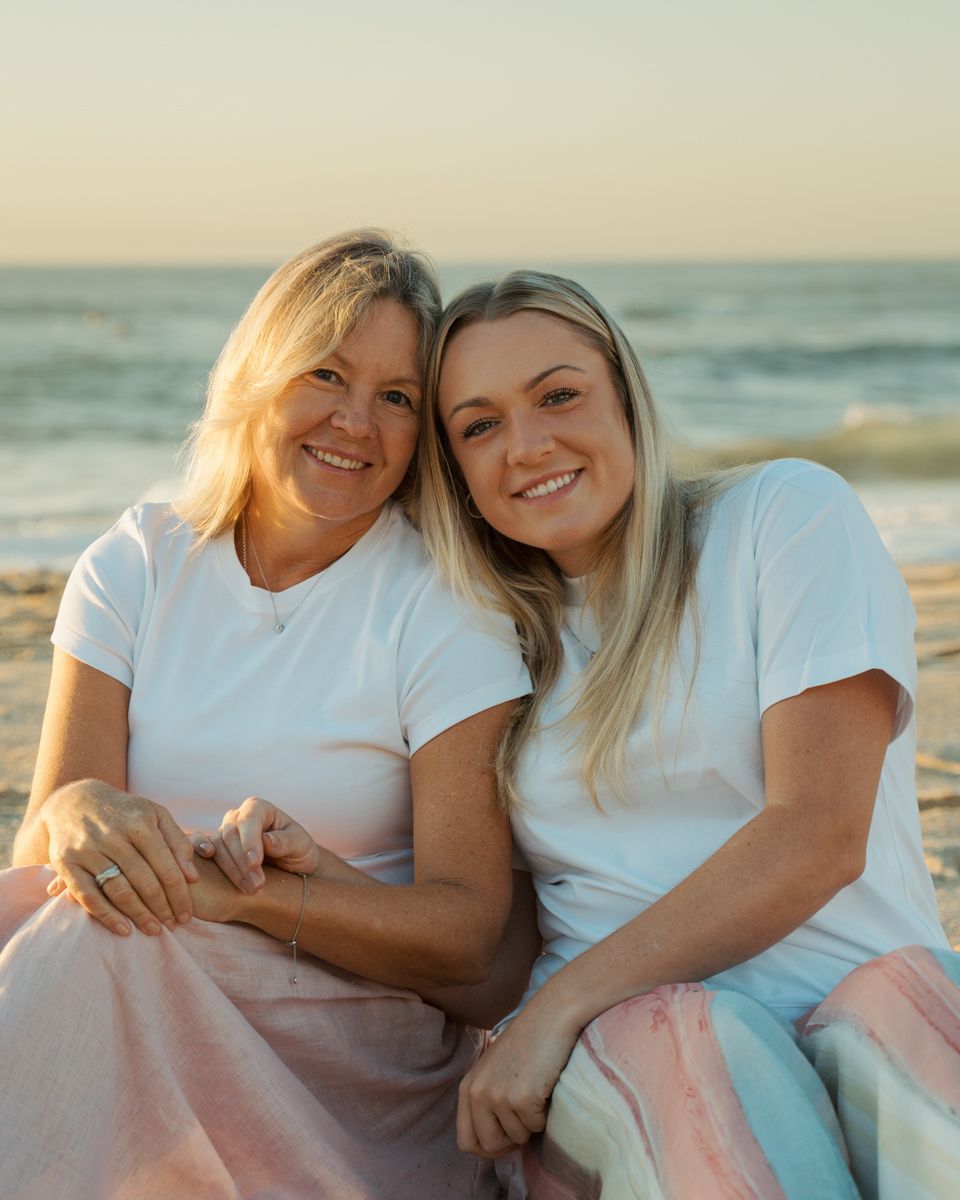 Two blonde women mom and daughter smiling together on a beach at sunrise, wearing matching white t-shirts and pastel skirts Bronte Beach