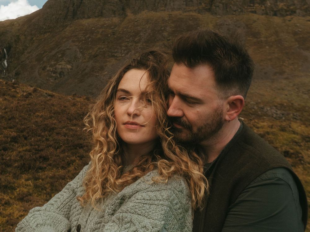 picture of couple in front of rugged green mountain, she's leaning against him and his eyes are closed