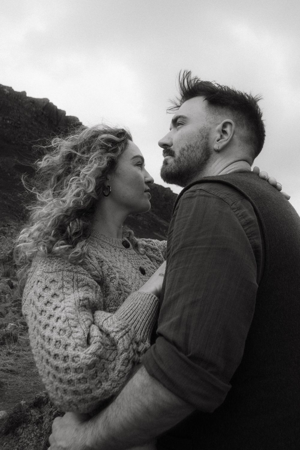 Black and white photo of Couple posing in rugged Irish nature, wind is picking up her hair as she's gazing past him