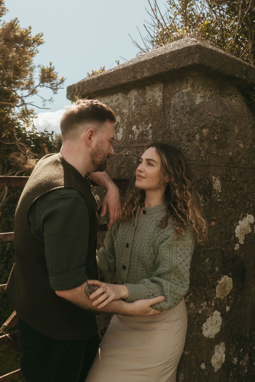 Couple gazing at each other leaning against a wall with trees in the background