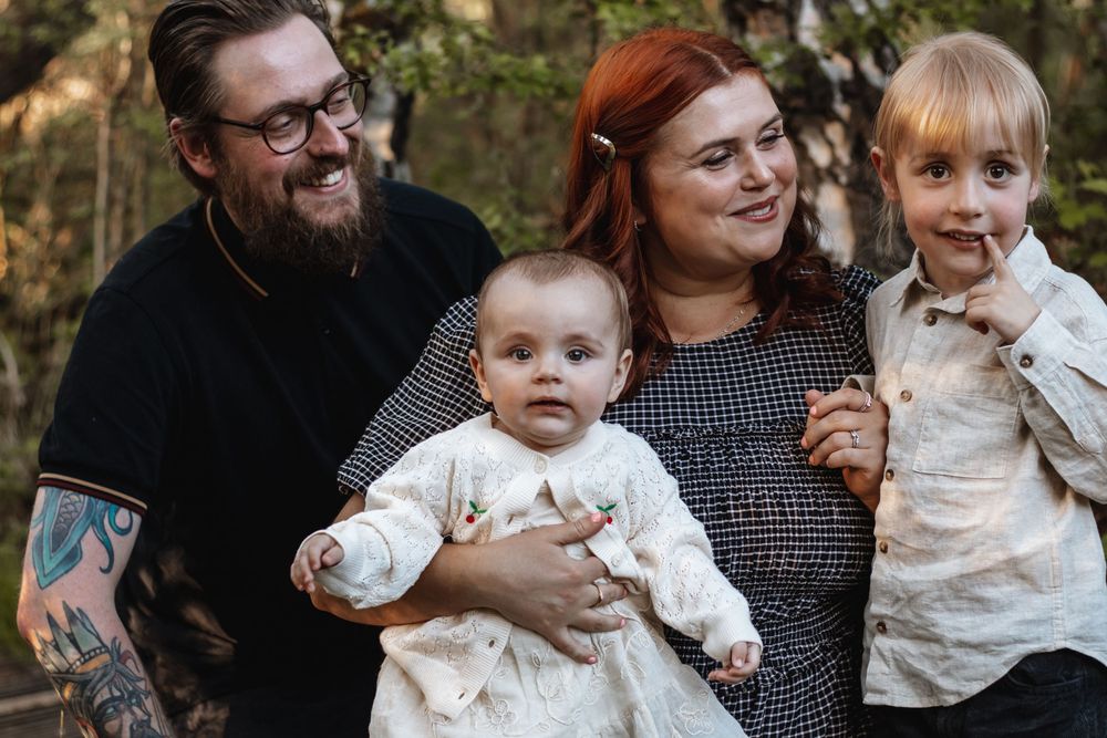 Happy family with tattooed dad, redhead mom, baby, and toddler smiling outdoors in a natural setting.