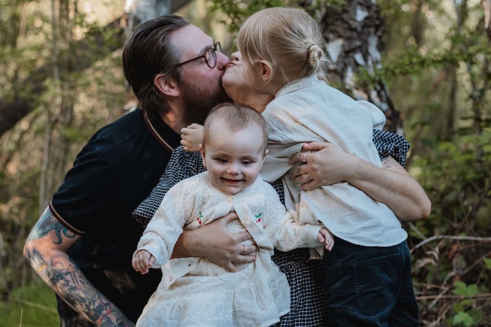 A tattooed father holds a smiling baby while a toddler kisses his cheek outdoors in a forest setting.
