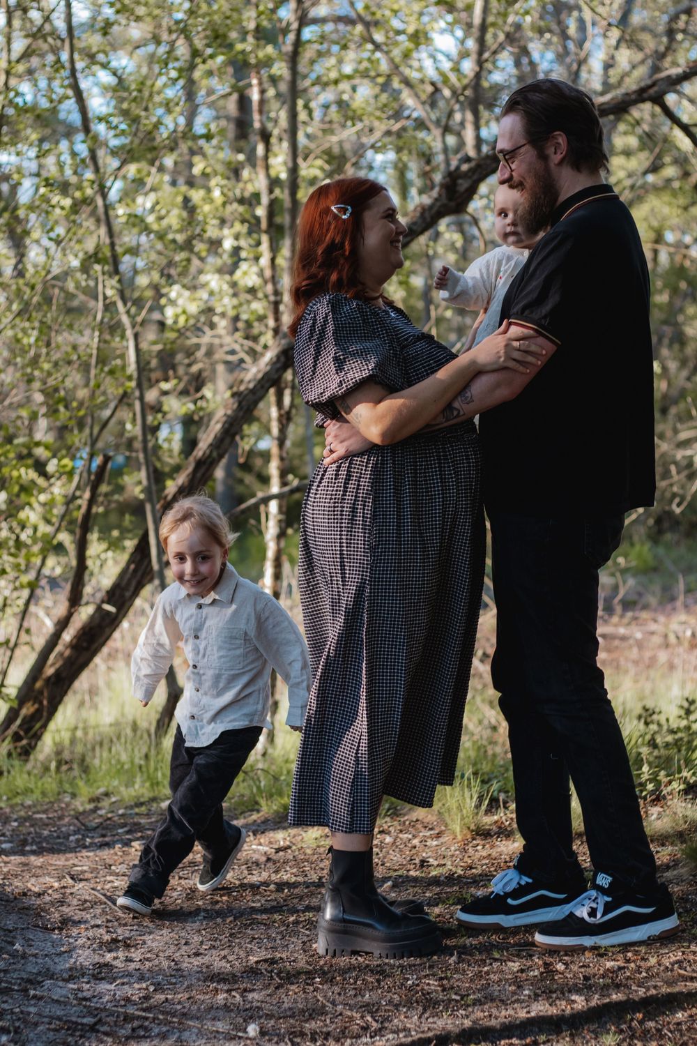 Pregnant woman and man embrace outdoors while two young children play around them in a sunlit forest setting.