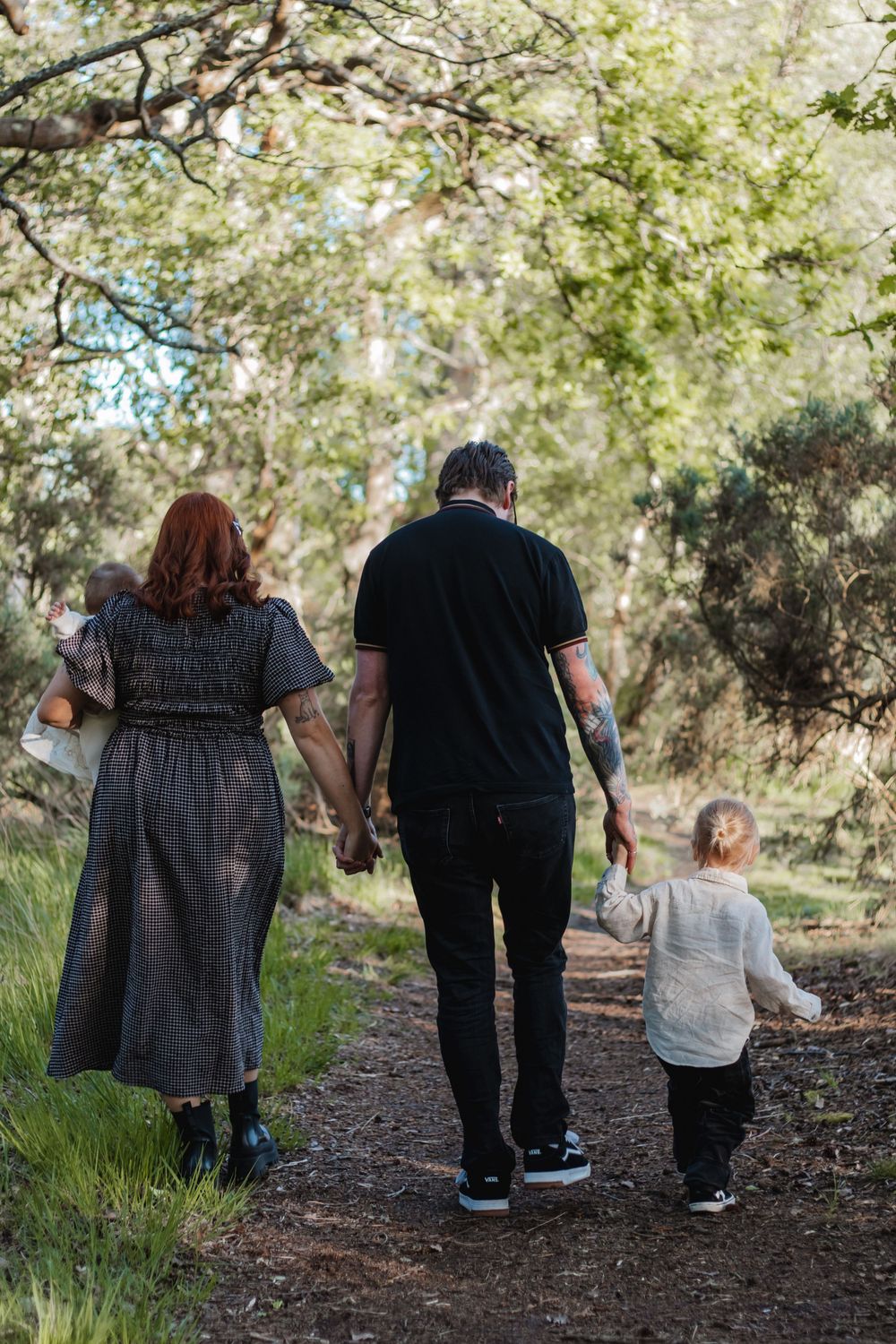 Family of four walking hand-in-hand down a scenic forest path, parents with tattooed arms and young child.