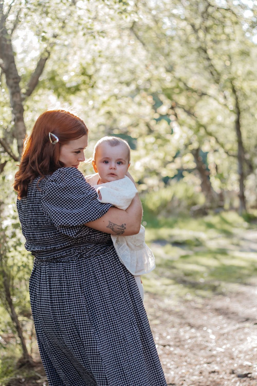 A redheaded mother in a plaid dress holds her baby outdoors in a sunlit green forest path.