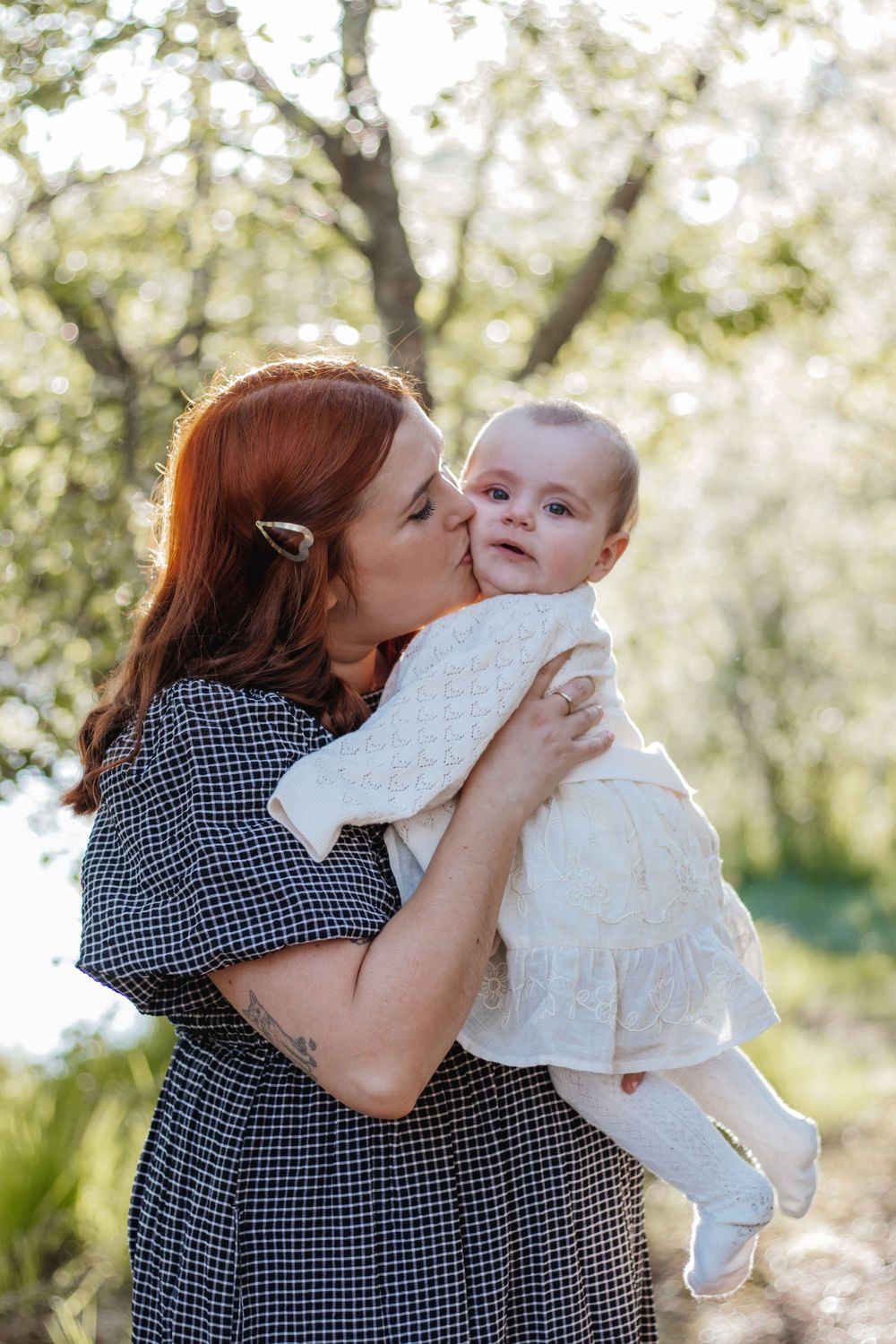 A red-haired mother kisses her smiling baby in a white dress outdoors among blooming trees in soft sunlight.