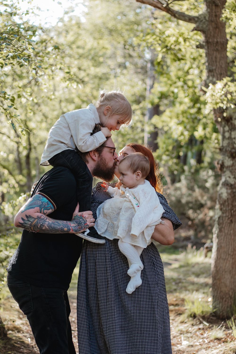 Tattooed father kisses baby while toddler sits on his shoulders outdoors in a sunlit forest.