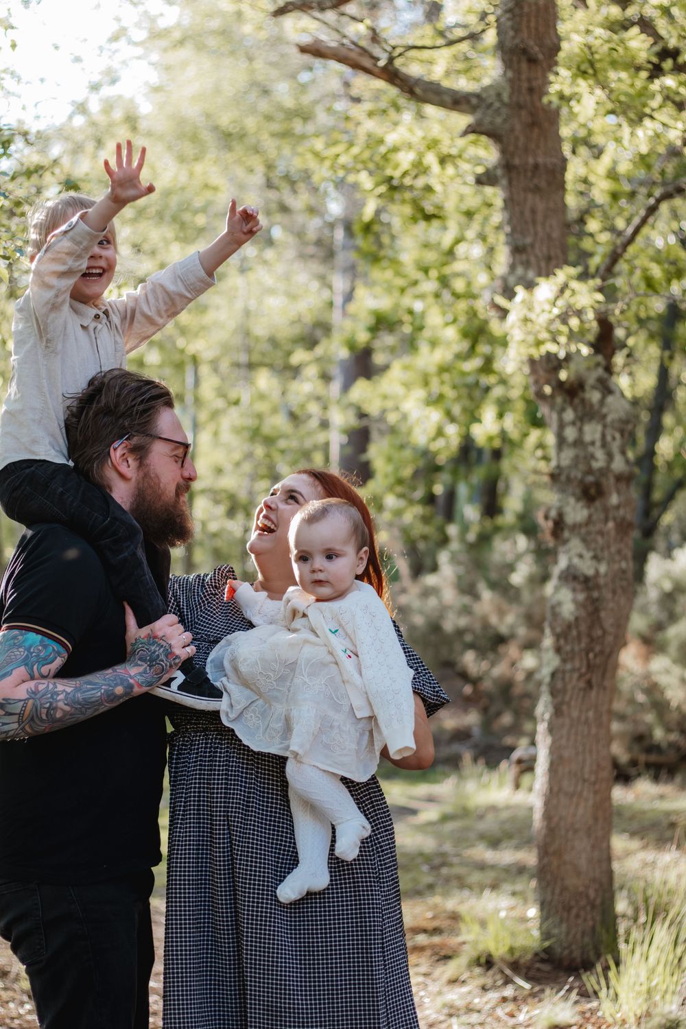 Joyful family with tattooed dad, redhead mom holding baby, and excited toddler on shoulders in forest.