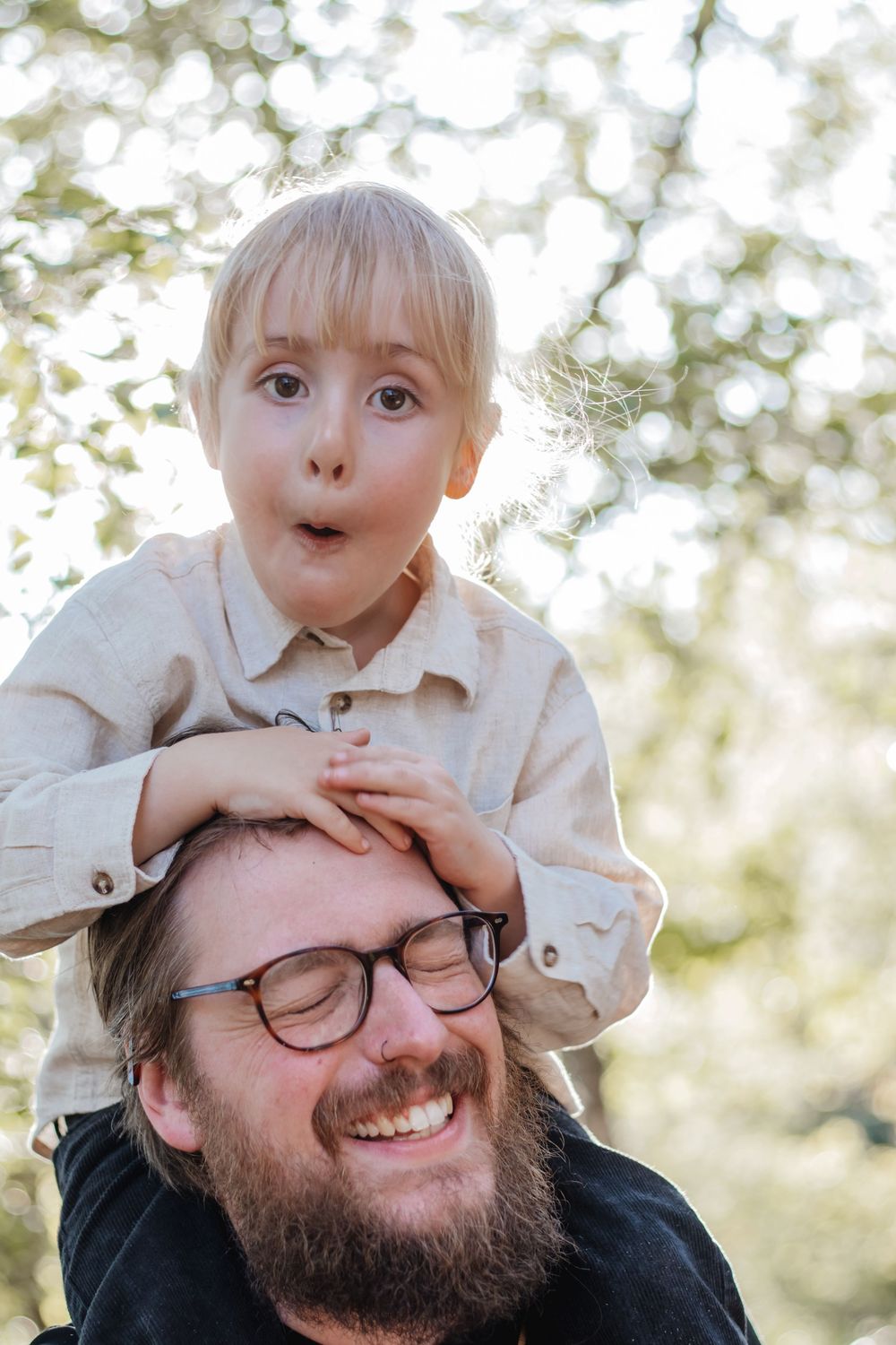 A laughing bearded man with glasses carries a wide-eyed blonde toddler on his shoulders outdoors.