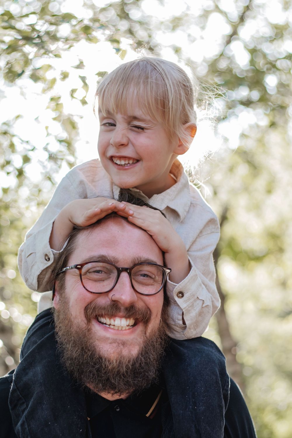 Smiling bearded father with glasses and laughing blonde toddler sitting on his shoulders outdoors.