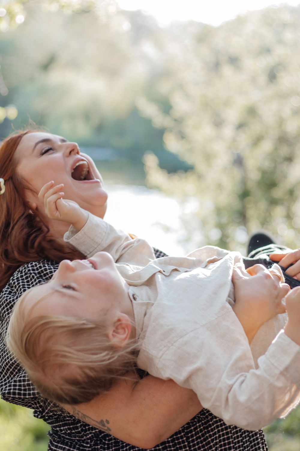 A joyful redhead mother laughs while playfully holding her blonde toddler outdoors in a sunny green park.