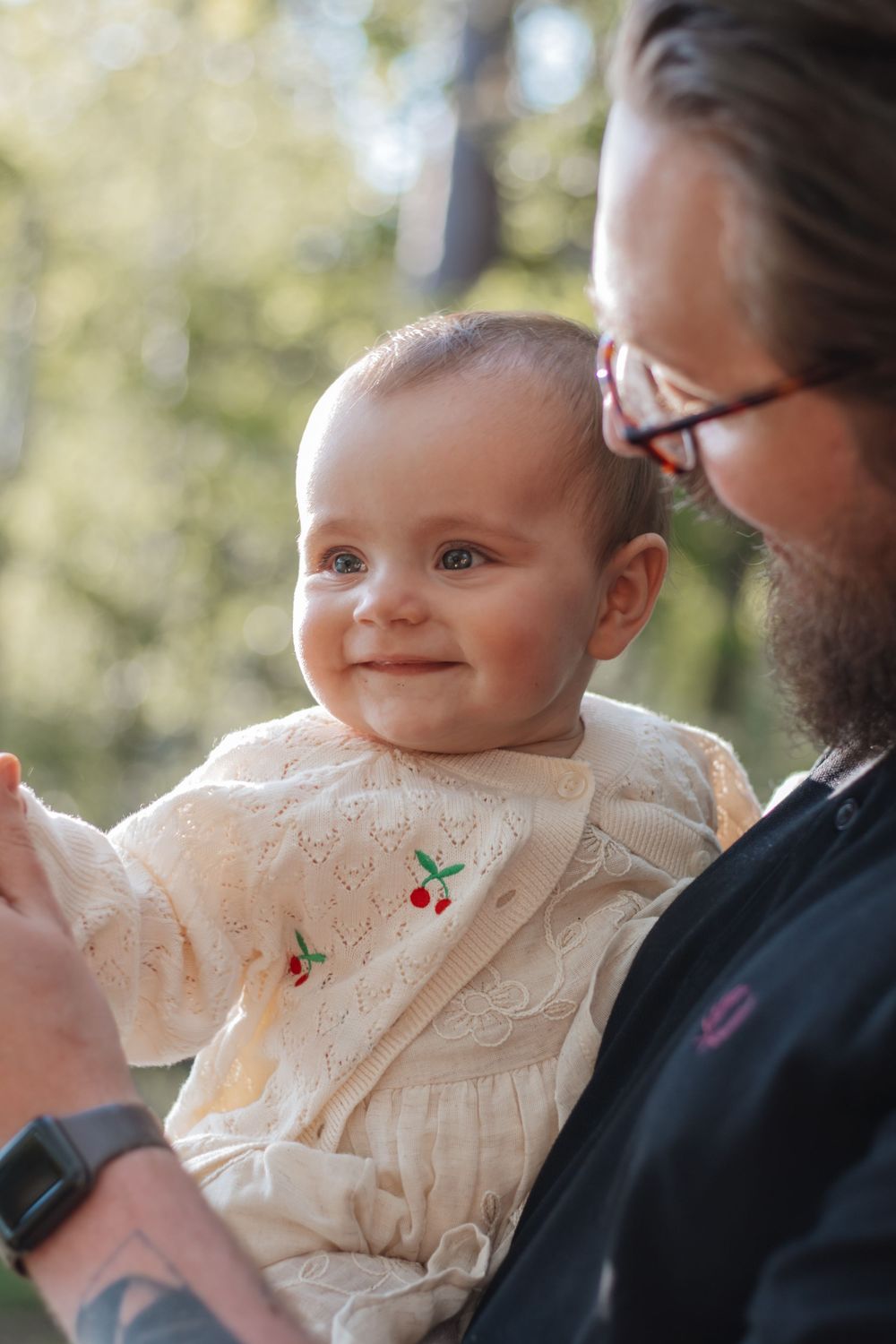 A smiling baby in a cherry-embroidered dress being held by a bearded man wearing glasses outdoors.