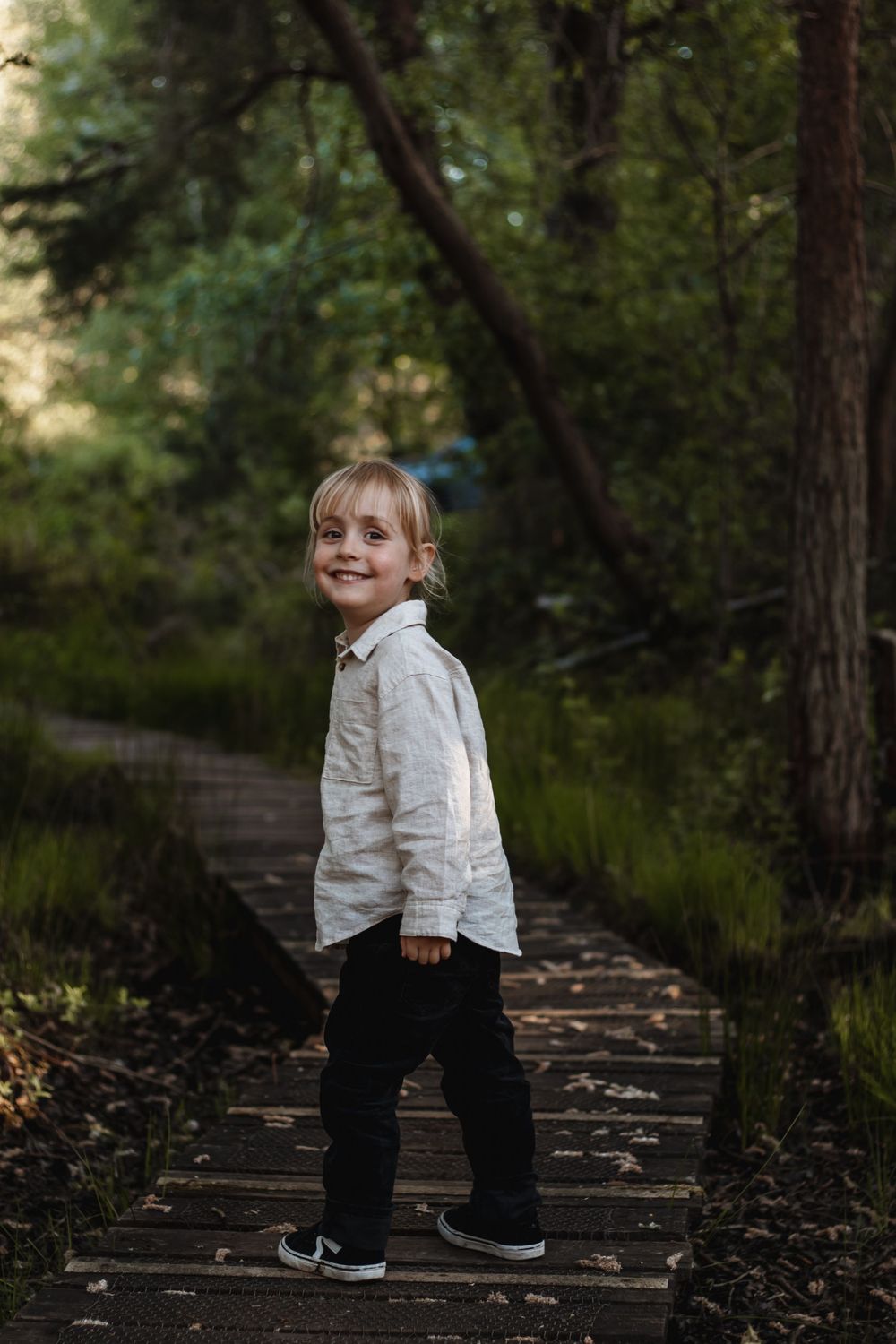 Smiling blonde child in white shirt and black pants standing on a wooden path in a lush green forest.