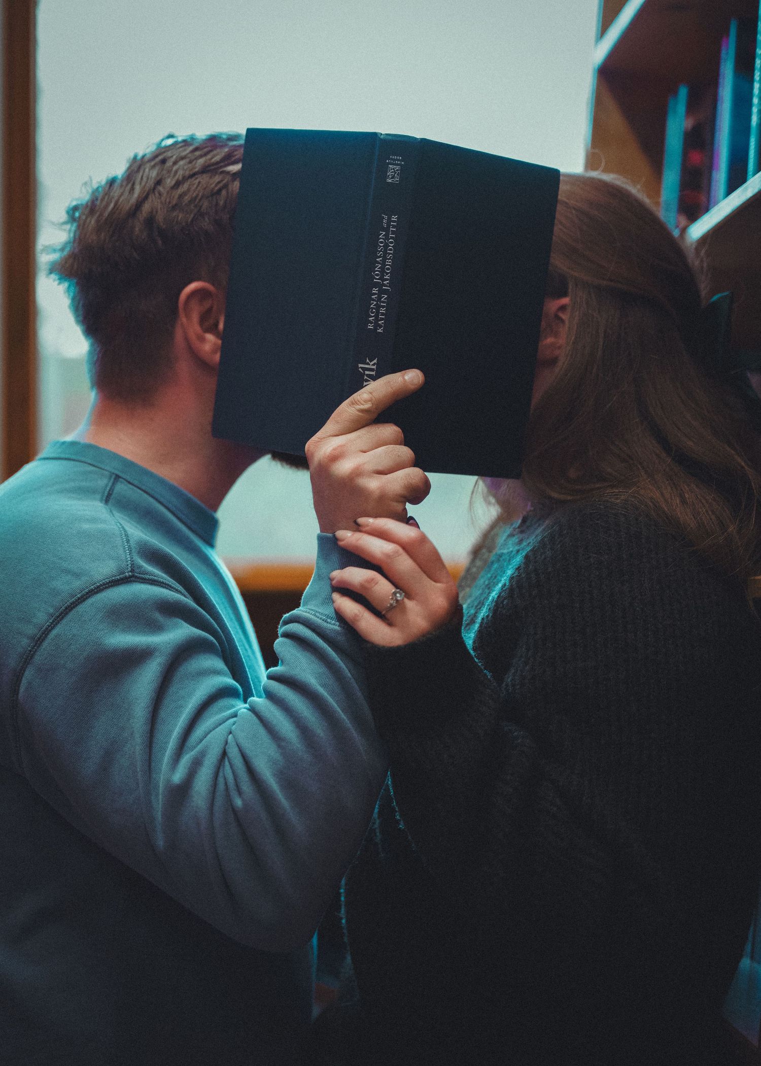 Two people hiding their faces behind a dark book in a library, appearing to share a secret kiss.