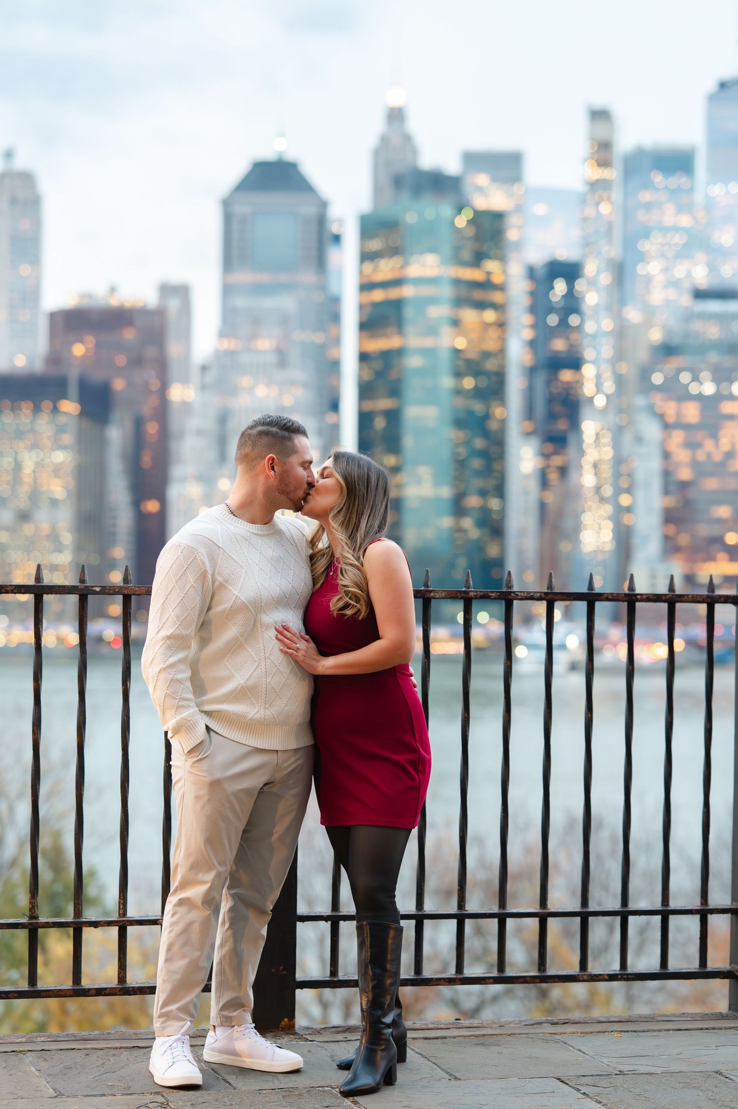 Couple kissing in front of NYC skyline at dusk, woman in red dress and boots, man in cream sweater and khakis.