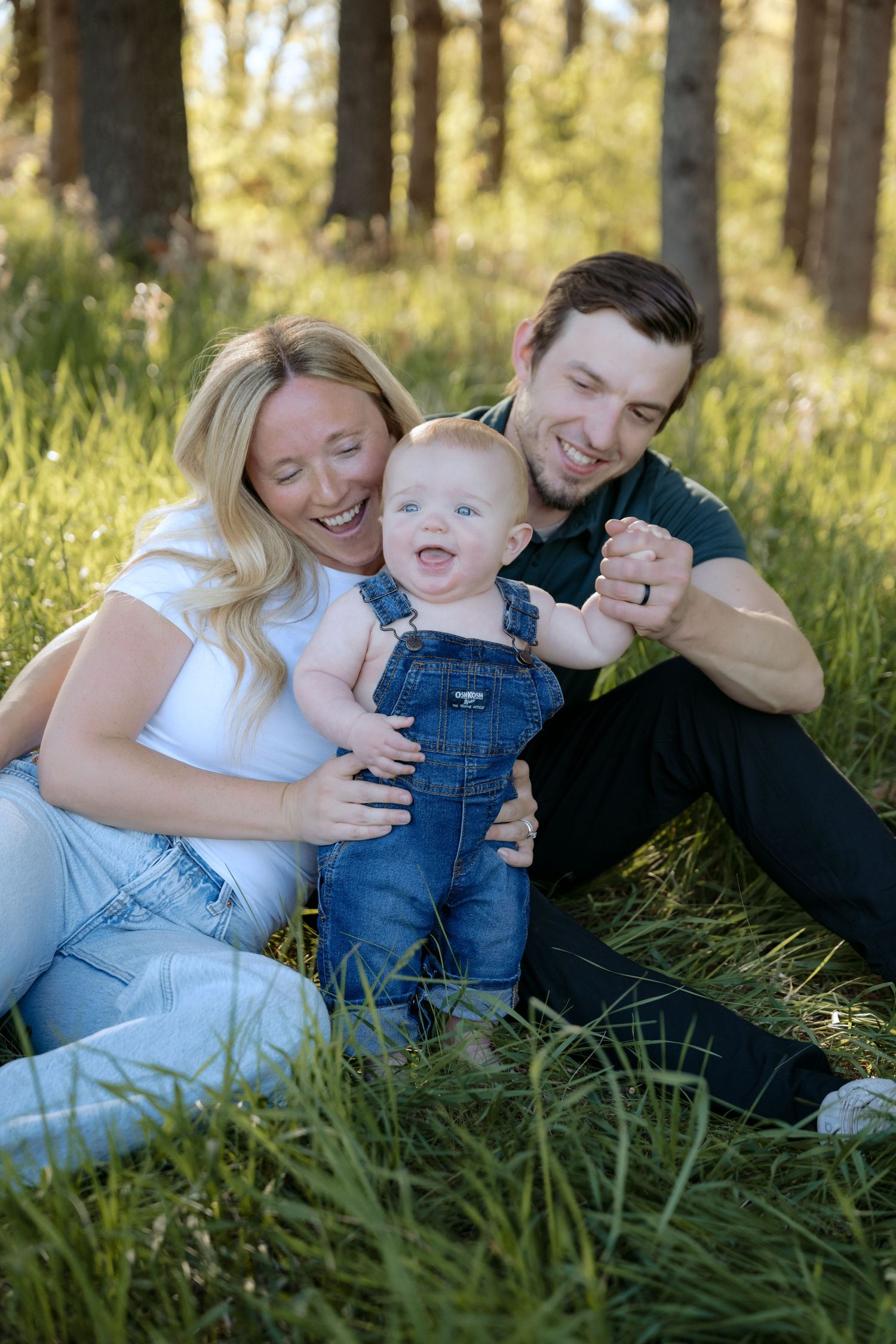 mom, dad, and baby boy in overalls sit in the sunlit grass for a spring outdoor family session at WI parks in hudson, wisconsin