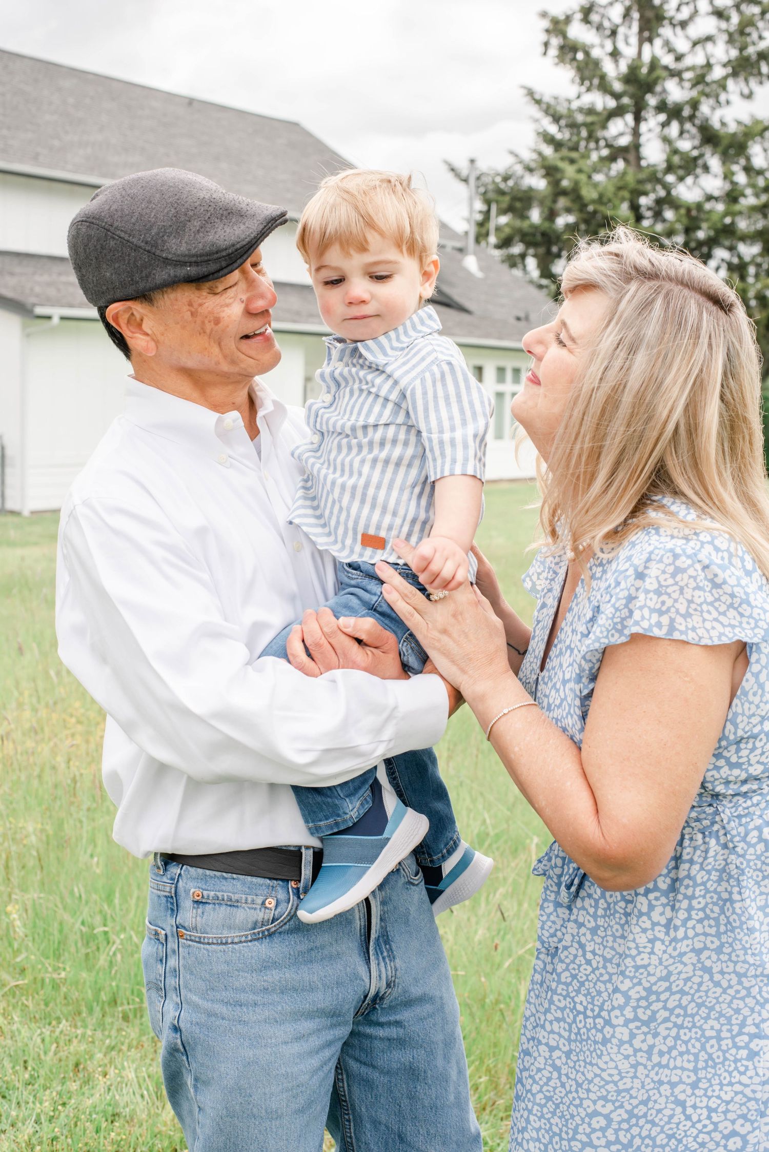 Grandparents smiling and holding a toddler boy outdoors on a green lawn near a white house.