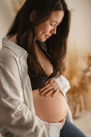 Pregnant woman in white shirt and black lace bra gently cradling her baby bump, looking down with a serene smile.