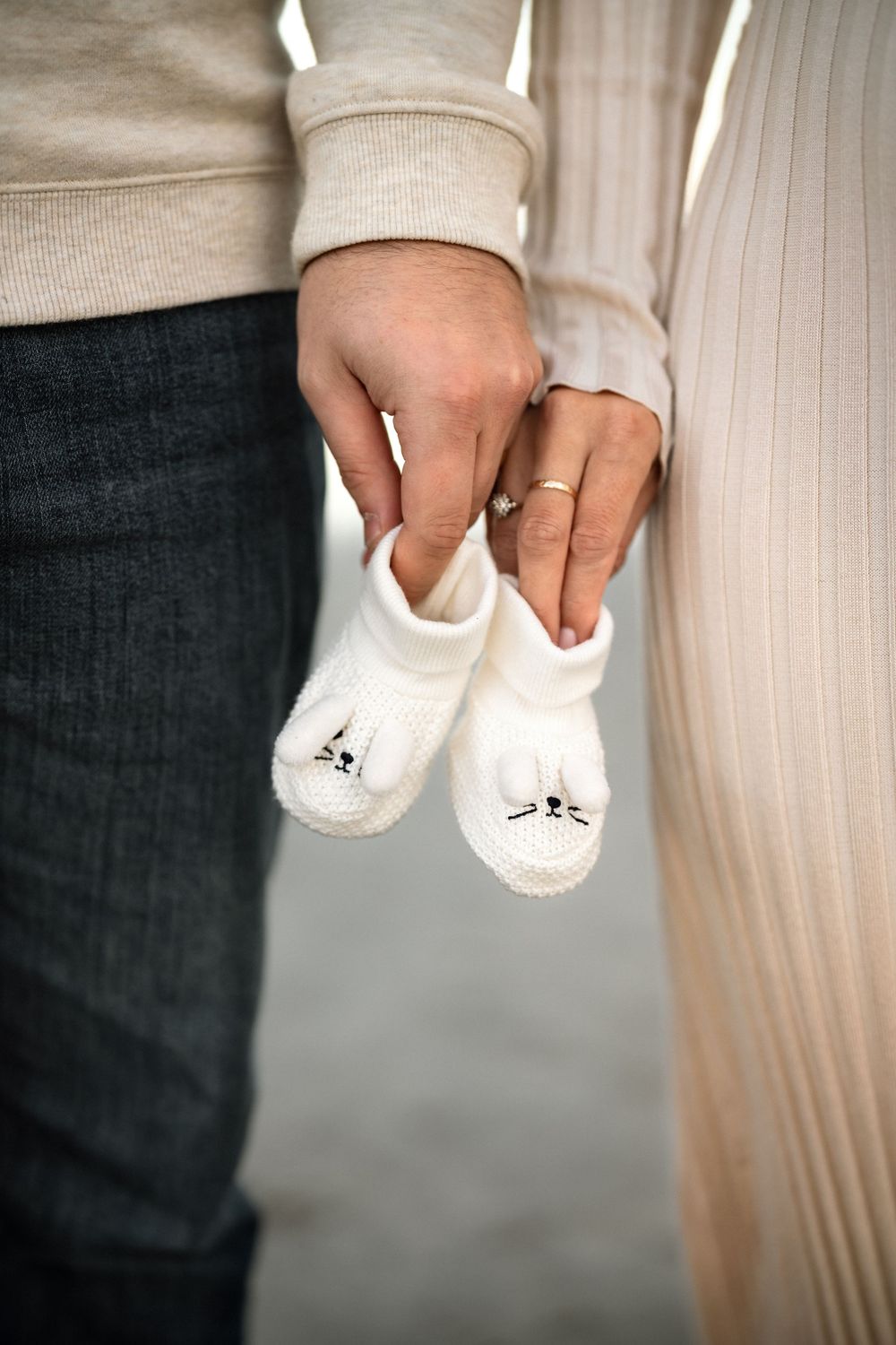 Couple holding tiny white knitted baby booties with cat faces, announcing pregnancy, woman wearing gold ring.