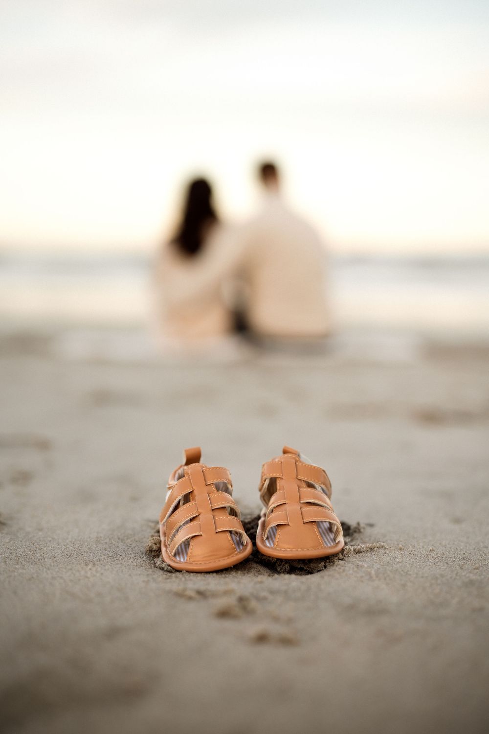 Small baby sandals in beach sand with blurred couple sitting by ocean in background at sunset.