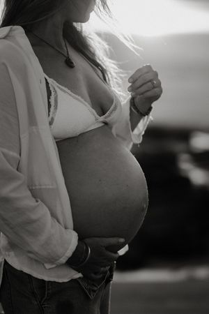 Black and white photo of a pregnant woman cradling her baby bump, wearing a bikini top and open hoodie outdoors.